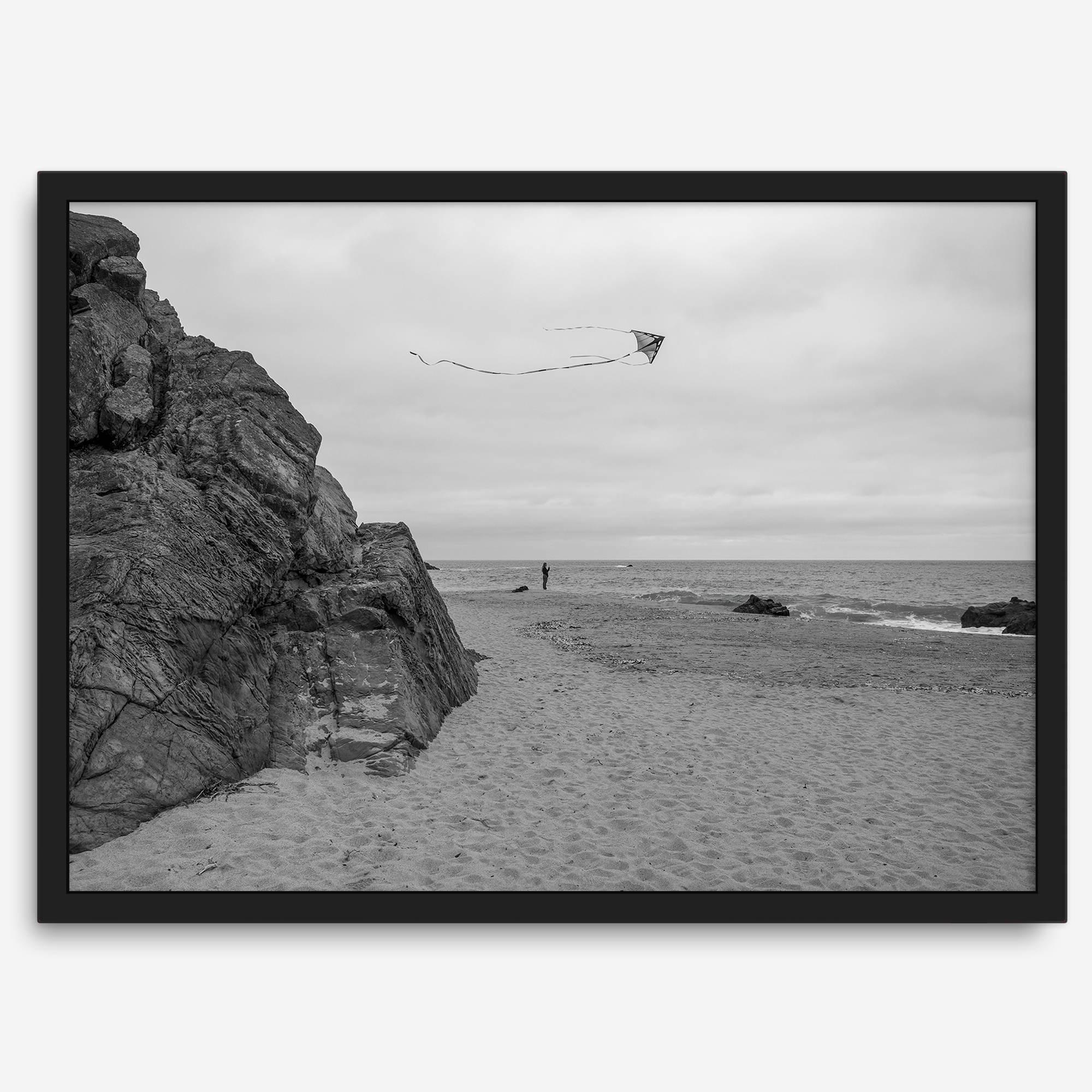 Black-and-white framed photo of a kite flying over a quiet California beach with rocky cliffs and an overcast sky.