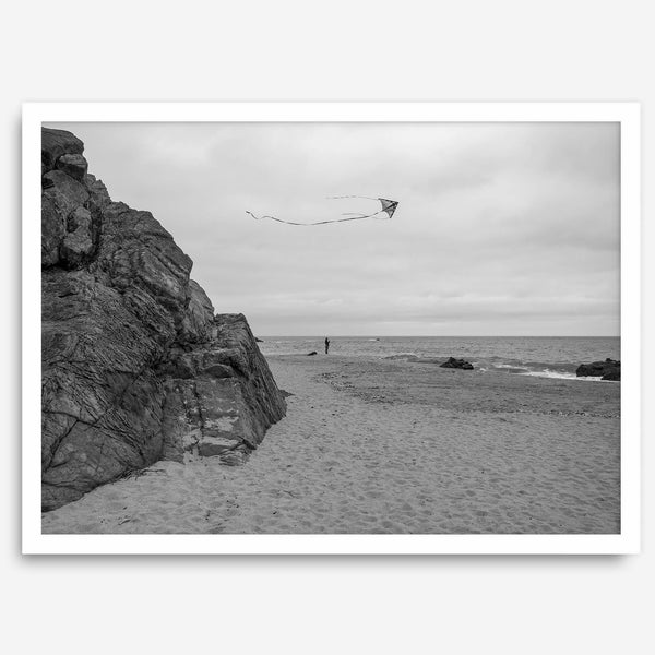 Black-and-white framed photo of a kite flying over a quiet California beach with rocky cliffs and an overcast sky.