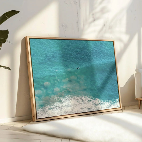 Aerial ocean canvas print showing a lone surfer paddling through turquoise waves with white surf along the beach shoreline