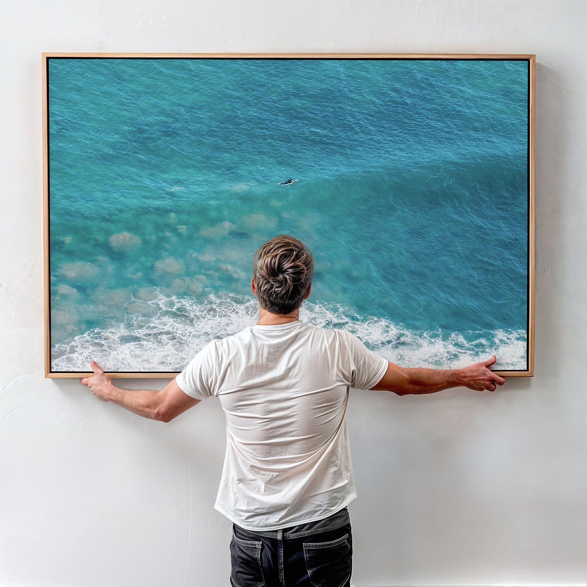 Aerial ocean canvas print showing a lone surfer paddling through turquoise waves with white surf along the beach shoreline