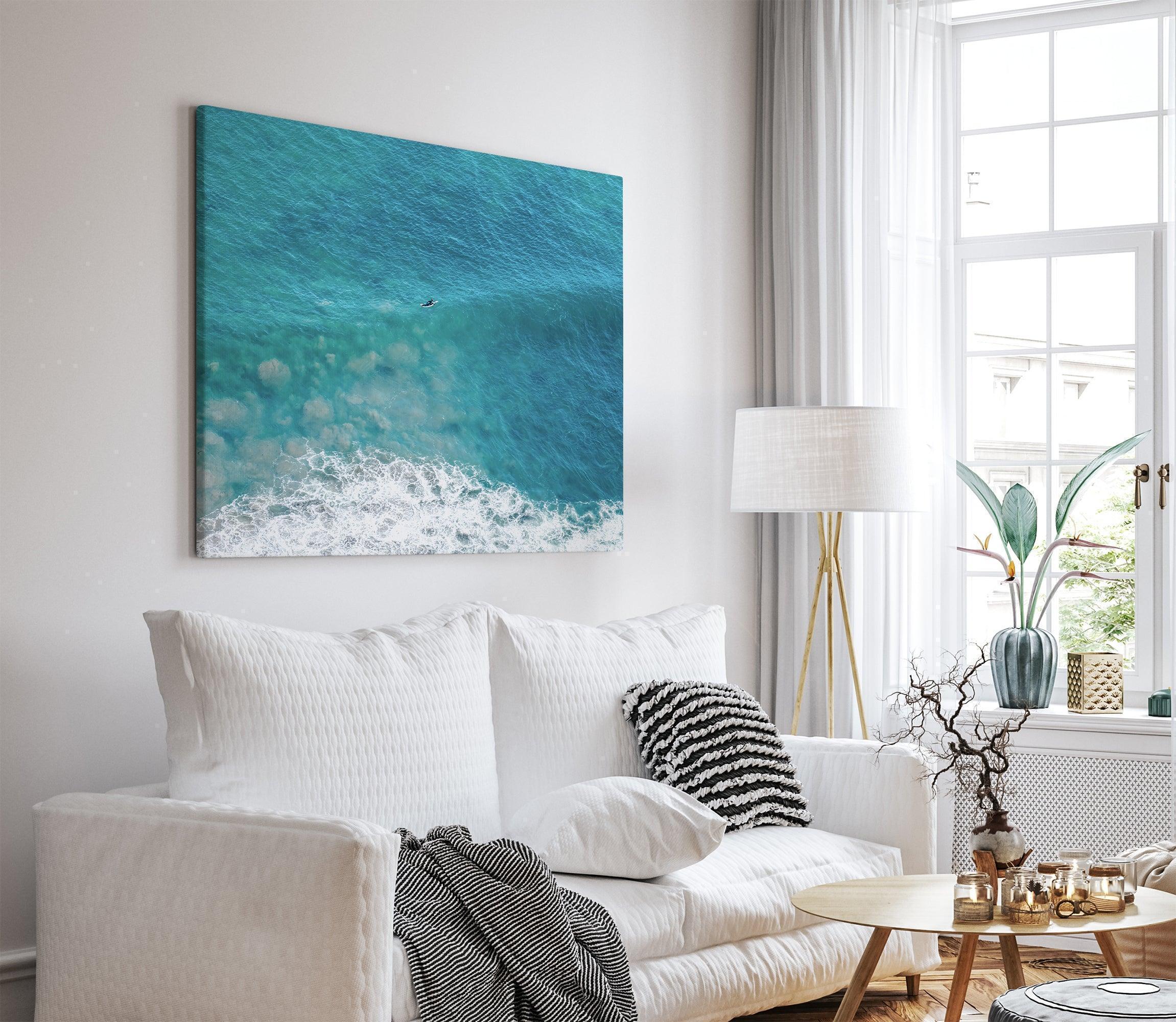 Aerial ocean canvas print showing a lone surfer paddling through turquoise waves with white surf along the beach shoreline