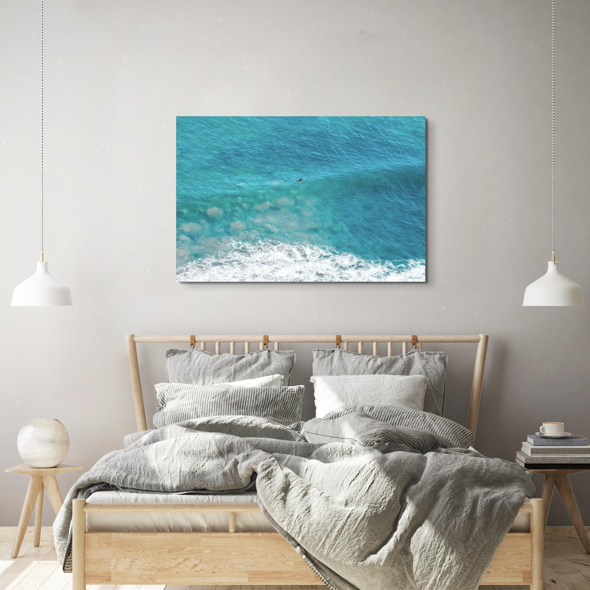 Aerial ocean canvas print showing a lone surfer paddling through turquoise waves with white surf along the beach shoreline