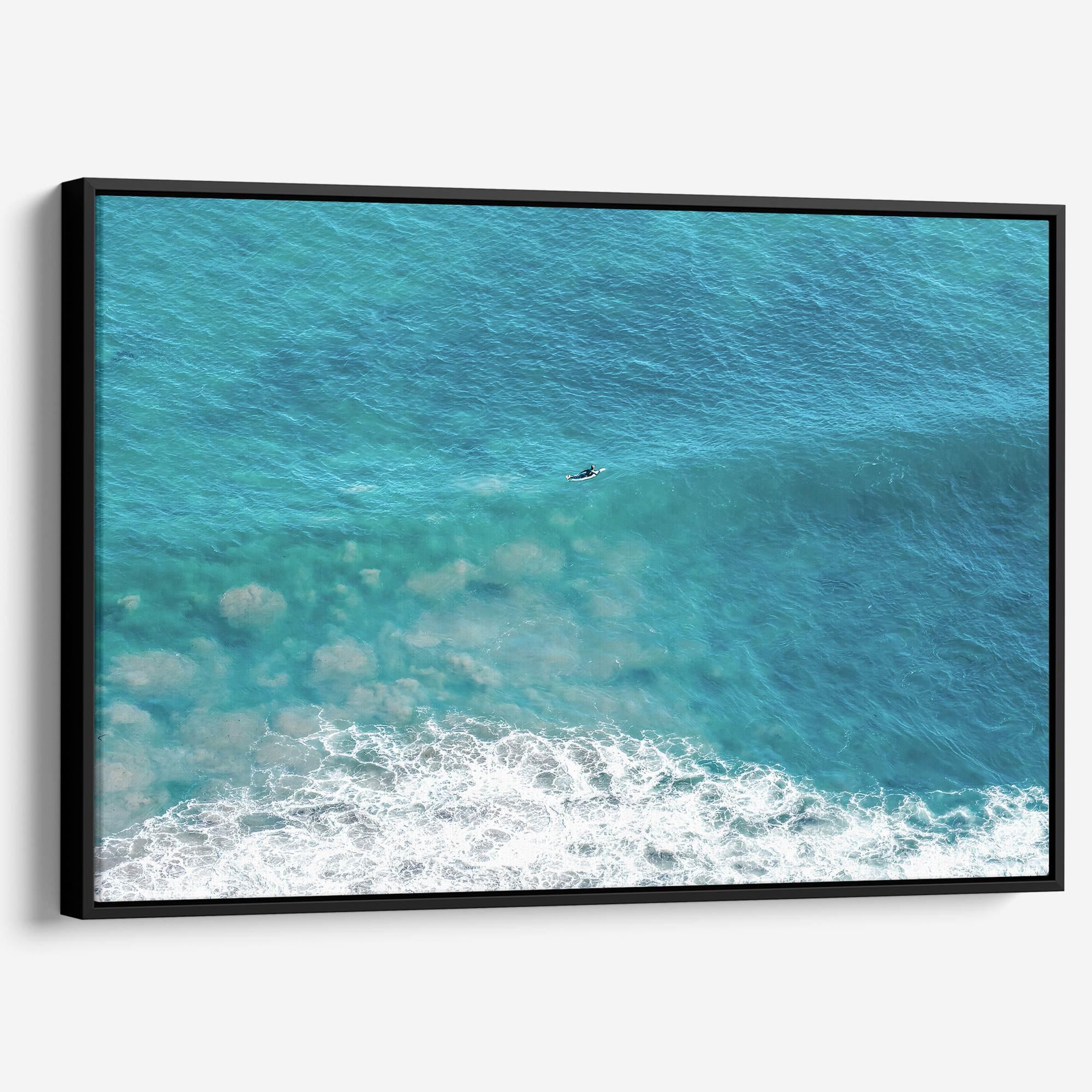 Aerial ocean canvas print showing a lone surfer paddling through turquoise waves with white surf along the beach shoreline