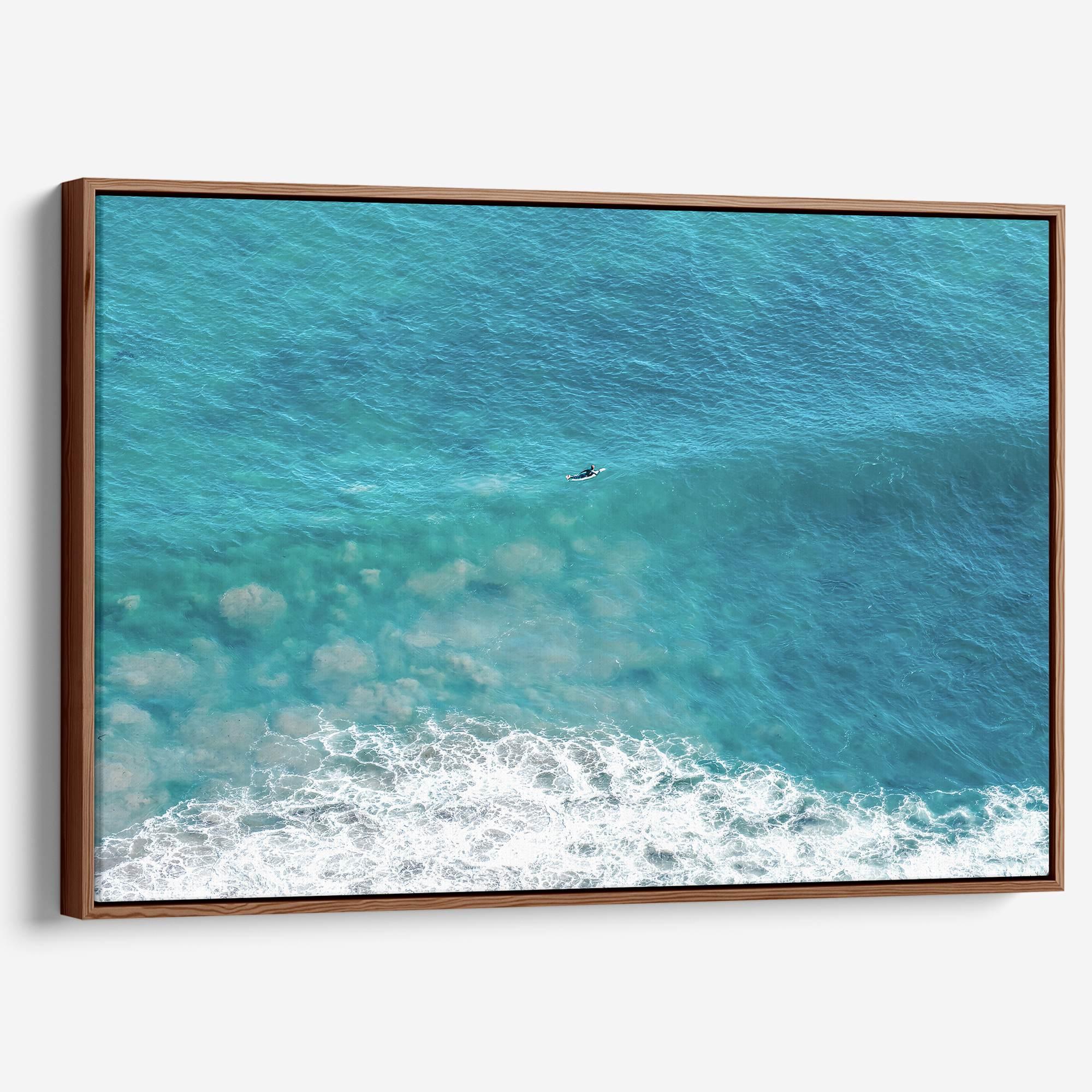 Aerial ocean canvas print showing a lone surfer paddling through turquoise waves with white surf along the beach shoreline