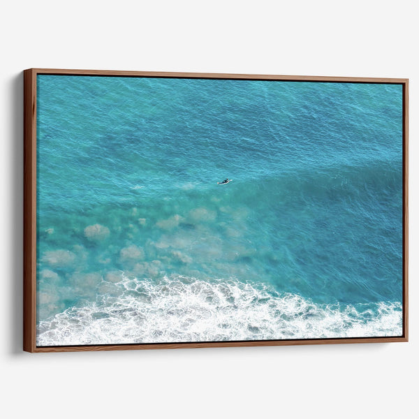 Aerial ocean canvas print showing a lone surfer paddling through turquoise waves with white surf along the beach shoreline