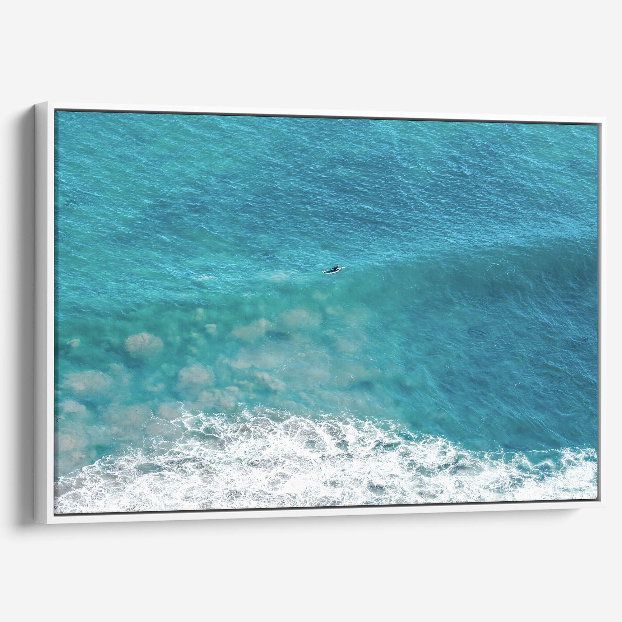 Aerial ocean canvas print showing a lone surfer paddling through turquoise waves with white surf along the beach shoreline