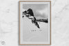 A fine art print showing five skiers traversing a vast, snowy mountain landscape under a pale winter sky, surrounded by untouched wilderness.
