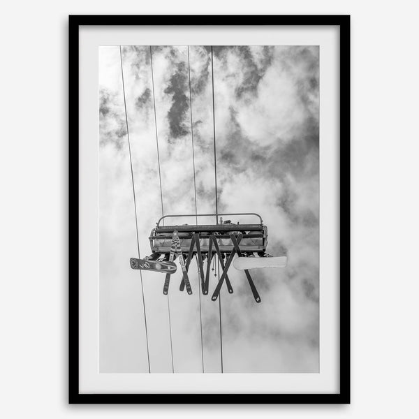 Monochrome ski lift view from below with hanging skis and cloudy mountain sky background.