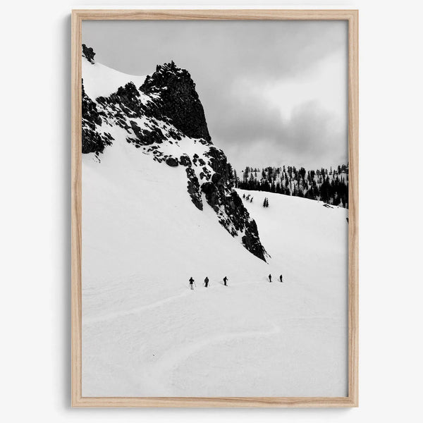 A fine art print showing five skiers traversing a vast, snowy mountain landscape under a pale winter sky, surrounded by untouched wilderness.