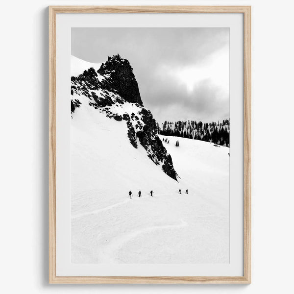 A fine art print showing five skiers traversing a vast, snowy mountain landscape under a pale winter sky, surrounded by untouched wilderness.
