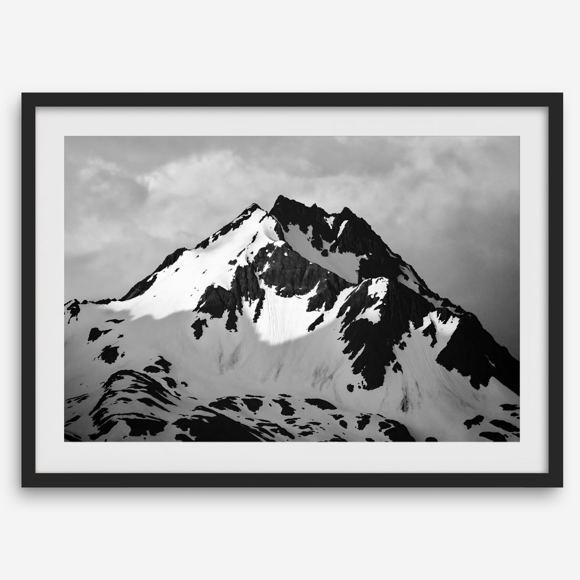 Black and white fine art photograph of a snowy Alaska mountain rising above coastal waters beneath dramatic clouds.