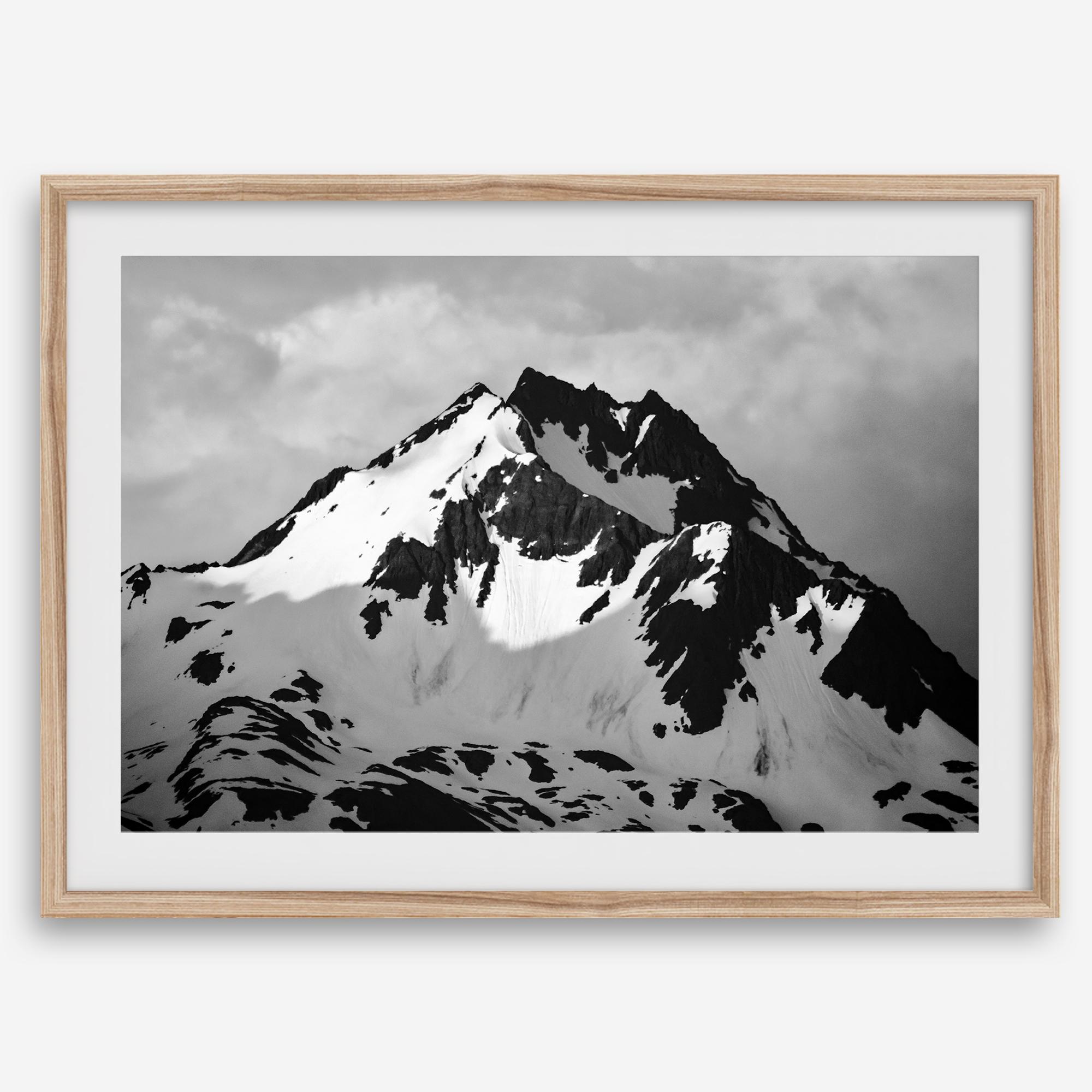 Black and white fine art photograph of a snowy Alaska mountain rising above coastal waters beneath dramatic clouds.