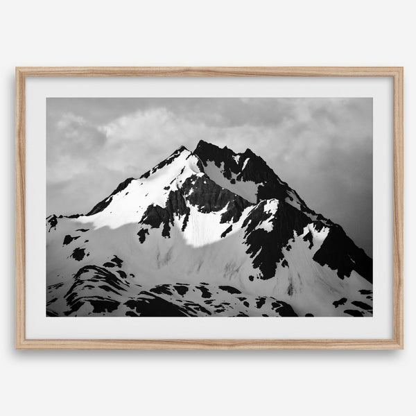 Black and white fine art photograph of a snowy Alaska mountain rising above coastal waters beneath dramatic clouds.