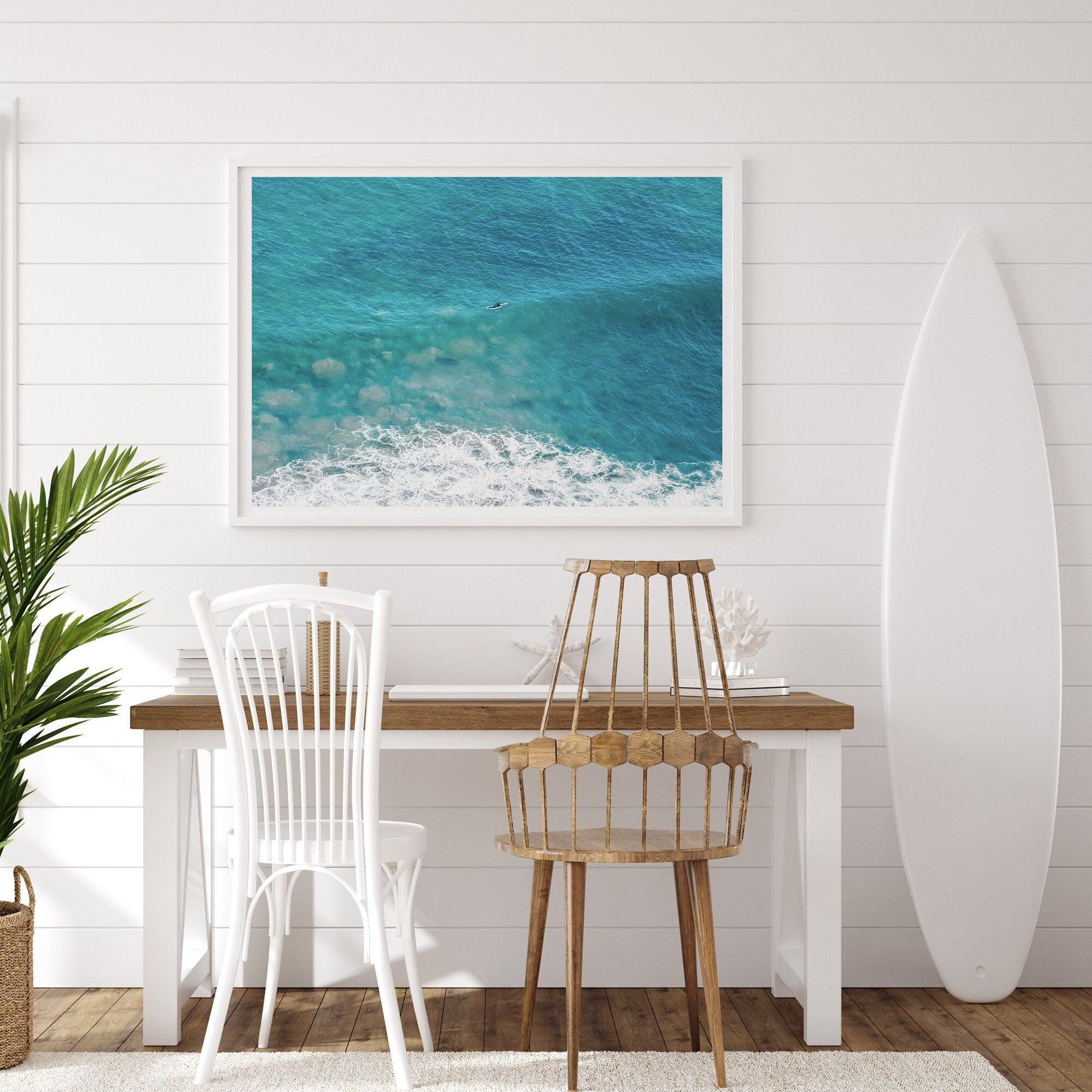 Aerial beach wall art featuring a lone surfer paddling through turquoise ocean waves, with foamy white surf along the shoreline.