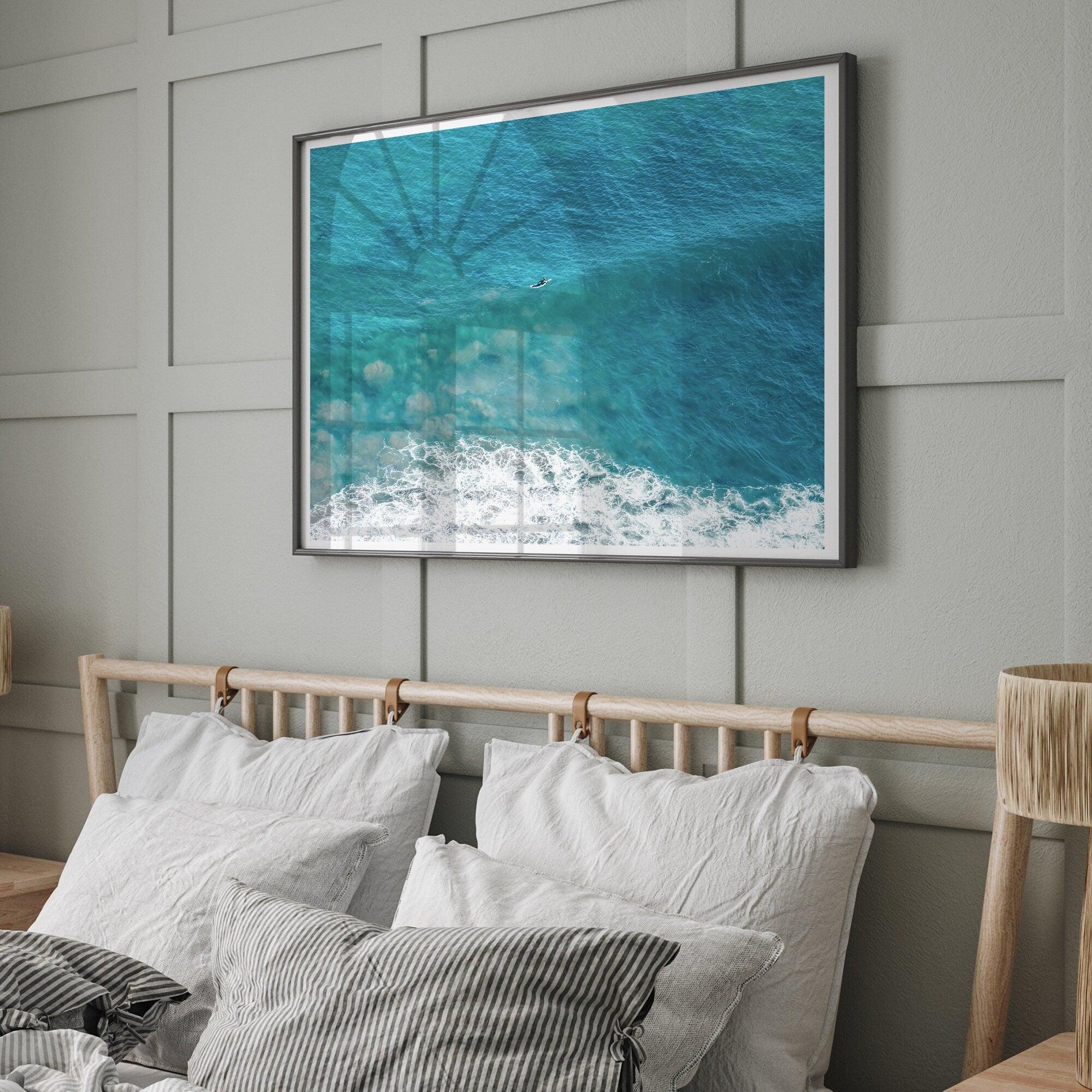 Aerial beach wall art featuring a lone surfer paddling through turquoise ocean waves, with foamy white surf along the shoreline.